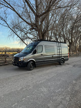 Grey Mercedes Sprinter adventure van on a gravel road, backed by bare trees. Ideal for RV rental getaways!