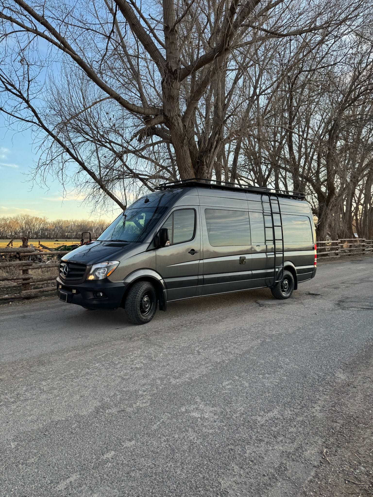 Grey Mercedes Sprinter adventure van on a gravel road, backed by bare trees. Ideal for RV rental getaways!