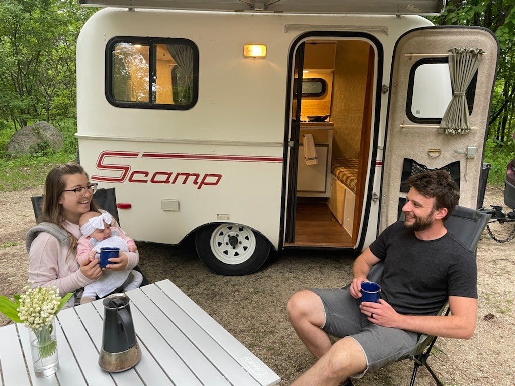 Smiling family with baby enjoys an RV rental experience, relaxing with coffee by their compact Scamp travel trailer.