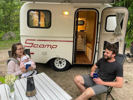 Smiling family with baby enjoys an RV rental experience, relaxing with coffee by their compact Scamp travel trailer.