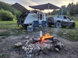 Relaxing RV rental campsite features a Bushwhacker teardrop trailer, truck, cooler, and a warm campfire at dusk.