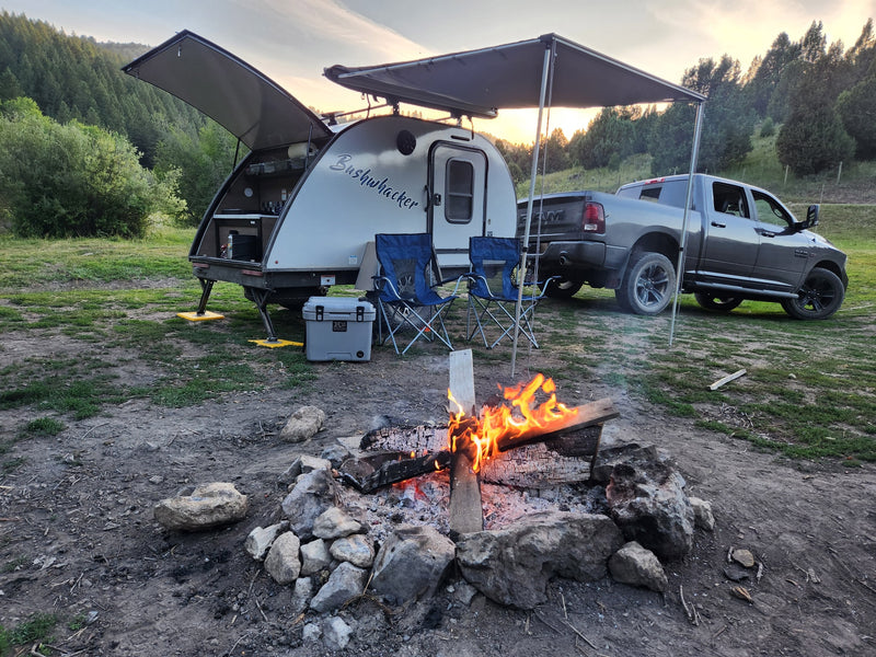 Relaxing RV rental campsite features a Bushwhacker teardrop trailer, truck, cooler, and a warm campfire at dusk. — photo 13