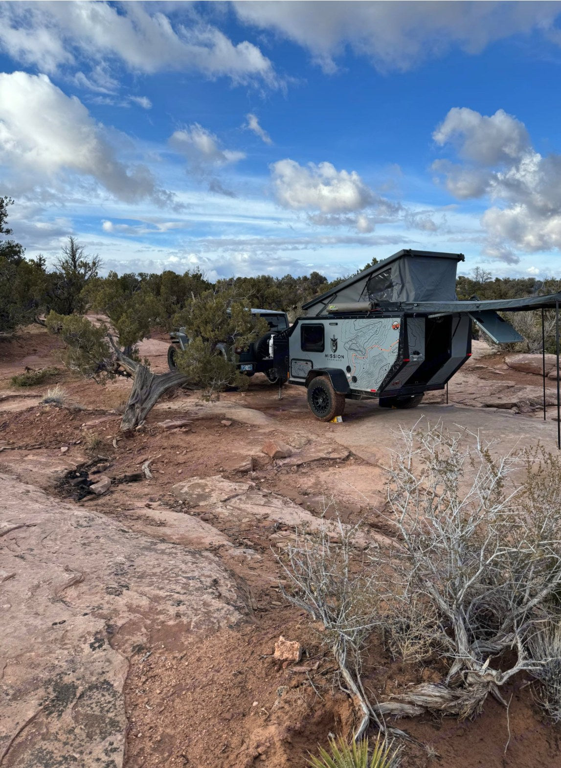 An off-road RV rental, a Mission Overland trailer with pop-up tent and awning, parked in a rugged red rock desert.