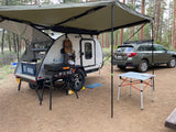 RV rental: A teardrop camper with its rear kitchen open, a grill, and an awning, parked next to a Subaru at a forest campsite.