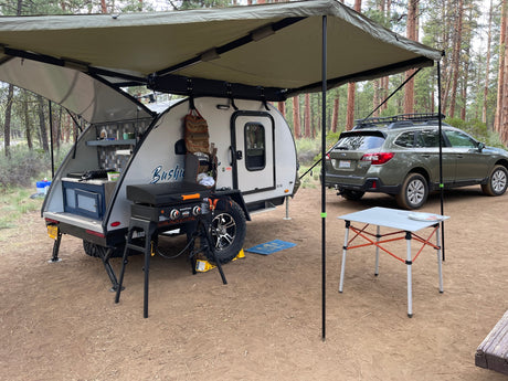 RV rental: A teardrop camper with its rear kitchen open, a grill, and an awning, parked next to a Subaru at a forest campsite.