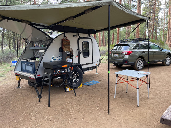 RV rental: A teardrop camper with its rear kitchen open, a grill, and an awning, parked next to a Subaru at a forest campsite.