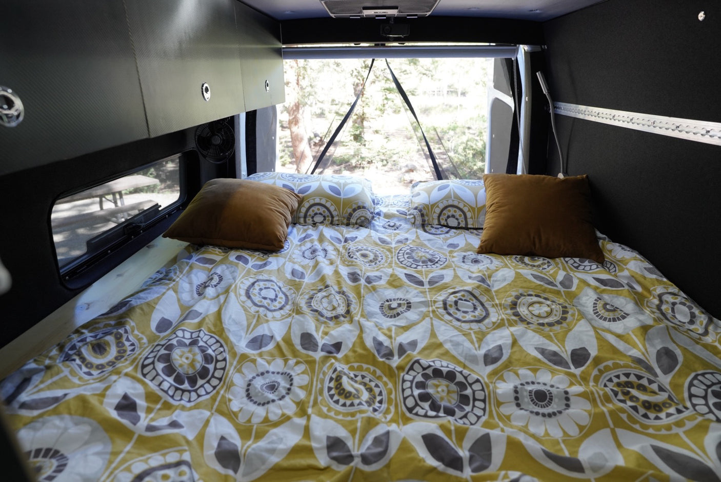 Cozy RV rental bed with yellow, white & grey patterned bedding, brown pillows, and a forest view. Perfect for camper travel.