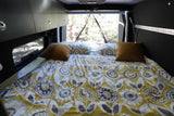 Cozy RV rental bed with yellow, white & grey patterned bedding, brown pillows, and a forest view. Perfect for camper travel.
