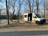 White RV rental van and picnic table by a peaceful lake, surrounded by bare trees on a gravel lot.
