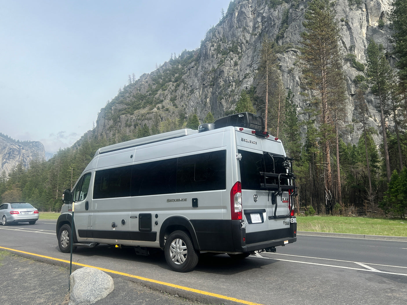 A grey Thor Sequence camper van (RV rental) with a bike rack is parked on a scenic road with large mountain cliffs and trees.