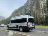 A grey Thor Sequence camper van (RV rental) with a bike rack is parked on a scenic road with large mountain cliffs and trees.