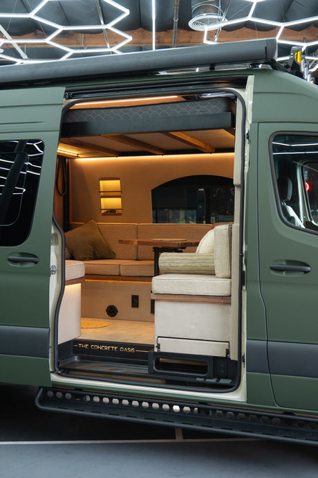 Inside an open RV rental van: a warm, modern custom interior with comfortable seating, table, and overhead sleeping area.