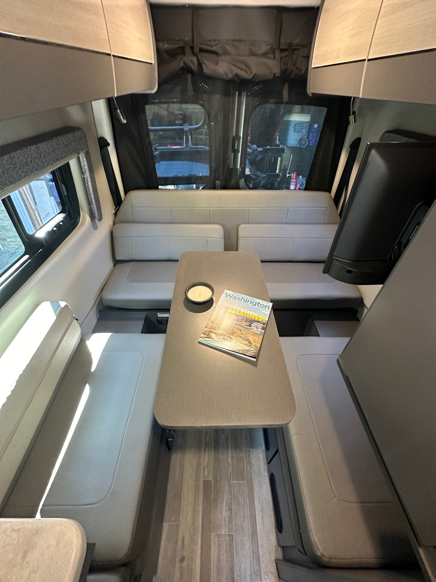 Cozy RV rental interior: dining area with benches, table, and TV. Overhead storage, windows offer outdoor view. Ideal for travel.
