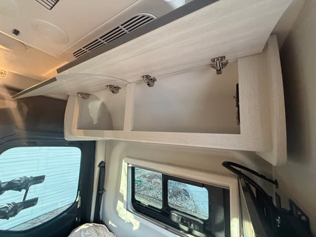 Inside an RV rental: Open overhead cabinets with light wood grain, ceiling vents, and side windows for storage and comfort.
