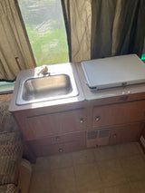 rv rental: A compact kitchenette with stainless steel sink, wood cabinets, and counter with white lid. Green outdoors visible.