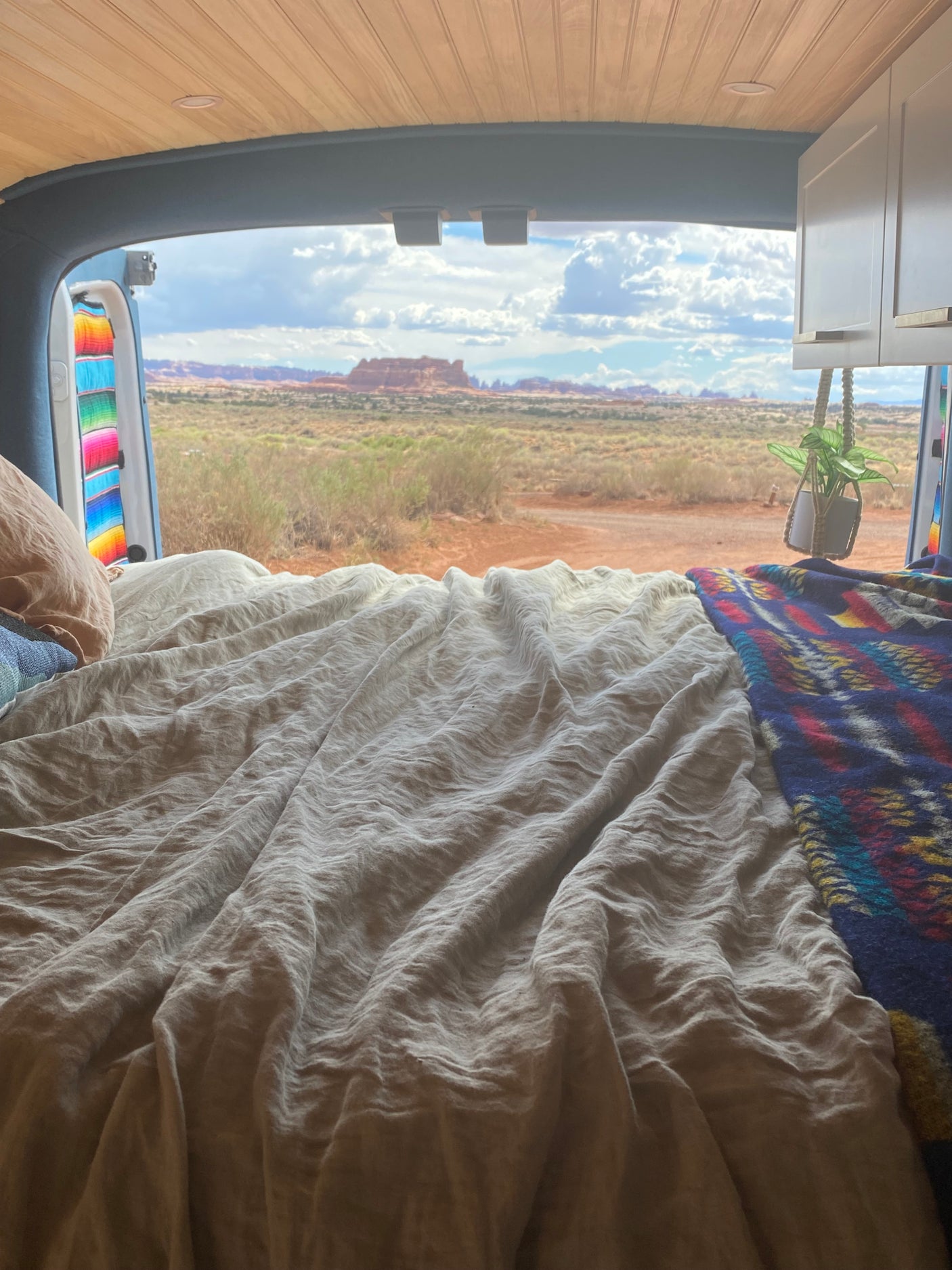 Inside an RV rental, a comfy bed faces open doors revealing a vast desert landscape with red rock formations.