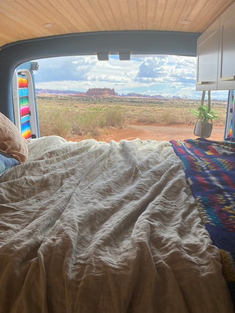 Inside an RV rental, a comfy bed faces open doors revealing a vast desert landscape with red rock formations.