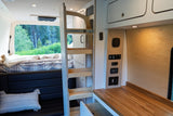This modern RV rental interior features a cozy bed with forest views, a functional wooden counter, and control panel, all within a well-designed camper van.
