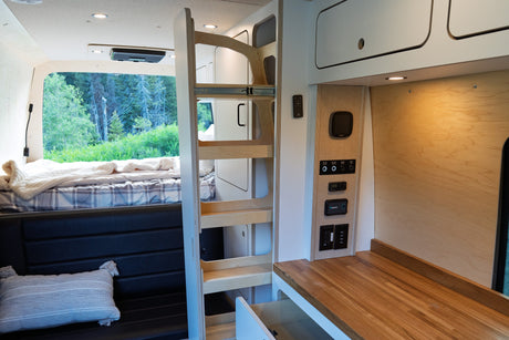 This modern RV rental interior features a cozy bed with forest views, a functional wooden counter, and control panel, all within a well-designed camper van.