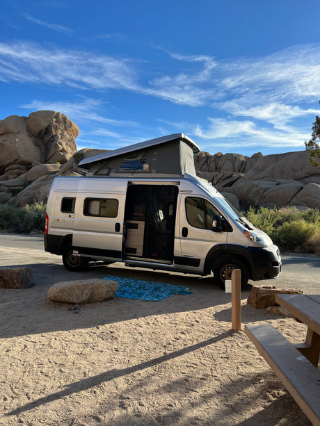 An RV rental pop-top camper van is parked at a sunny, rocky desert campsite. Its side door is open, revealing the interior.