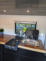 Modern RV rental kitchen with a 2-burner stove, sink, and wooden counters over dark cabinets, lit by a window.