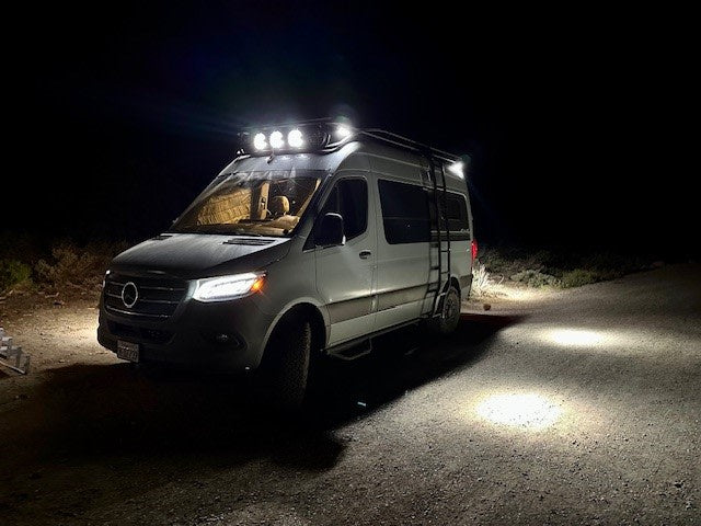 Brightly lit white Mercedes Sprinter van, a cozy rv rental, illuminating a dark dirt road at night with its powerful roof lights.