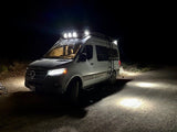 Brightly lit white Mercedes Sprinter van, a cozy rv rental, illuminating a dark dirt road at night with its powerful roof lights.