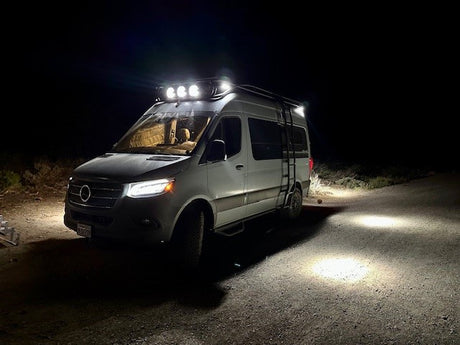 Brightly lit white Mercedes Sprinter van, a cozy rv rental, illuminating a dark dirt road at night with its powerful roof lights.