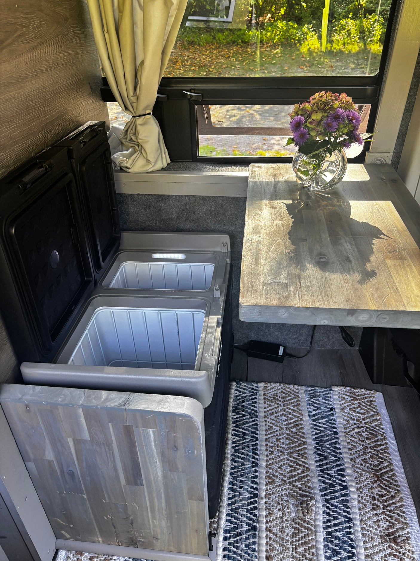 RV rental interior with open dual portable fridges, a wooden table holding flowers, and a window offering a lush view.