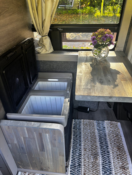 RV rental interior with open dual portable fridges, a wooden table holding flowers, and a window offering a lush view.