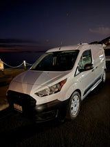 White Ford Transit Connect van, ideal for rv rental adventures, parked by the ocean at night under a starry sky.