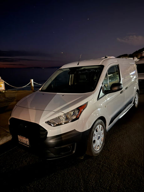 White Ford Transit Connect van, ideal for rv rental adventures, parked by the ocean at night under a starry sky.