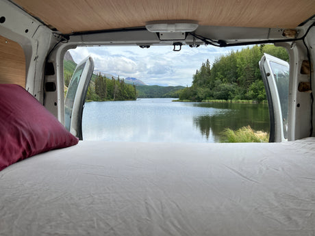 RV rental: Wake up to this serene lake and mountain view from a cozy bed in your van, doors open to nature.