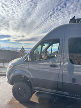 Off-road gray **RV rental** van with a dog peeking out, parked at a snowy mountain overlook.