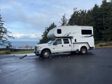 A large white truck camper, ideal for an RV rental, parked on asphalt overlooking the Pacific Coast. Evergreen trees frame the view.