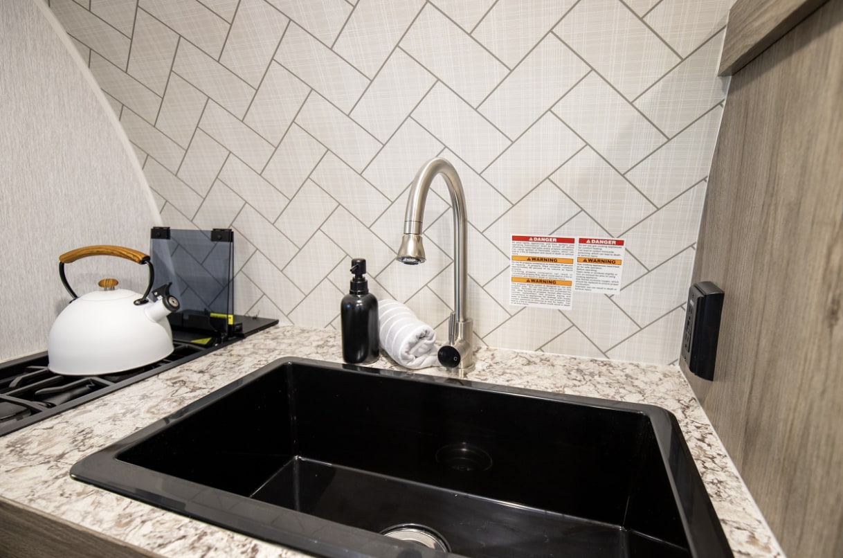 An RV rental's compact kitchen with a black sink, sleek faucet, herringbone patterned backsplash, and stovetop kettle.