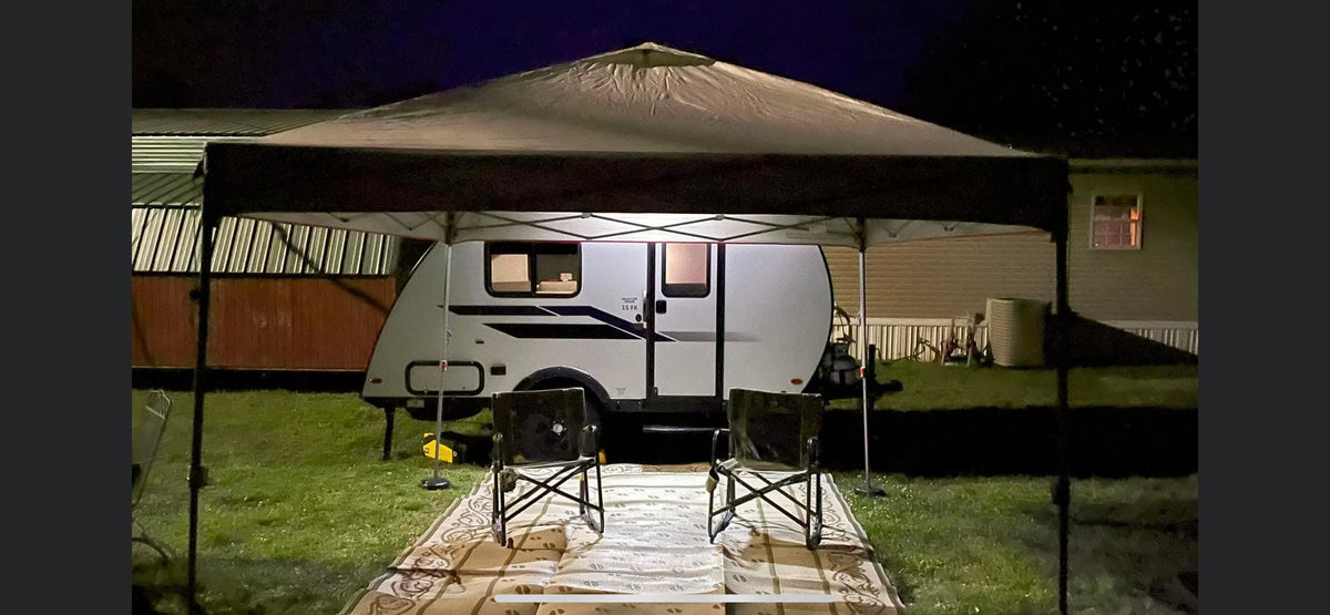 Cozy RV rental setup at night. A white travel trailer is under a lit canopy with two chairs on a rug