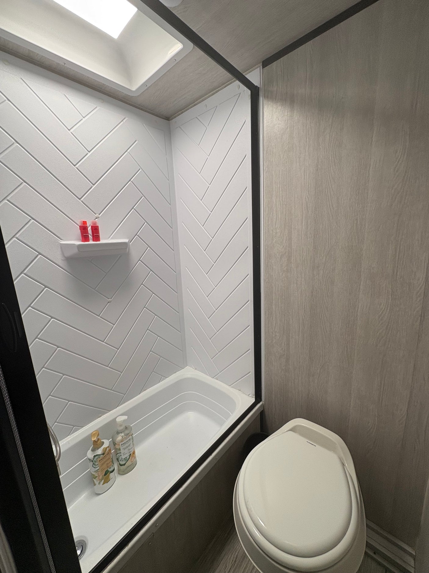 Clean RV rental bathroom with a white herringbone shower, bathtub, toilet, and wood-look walls. Features a skylight.