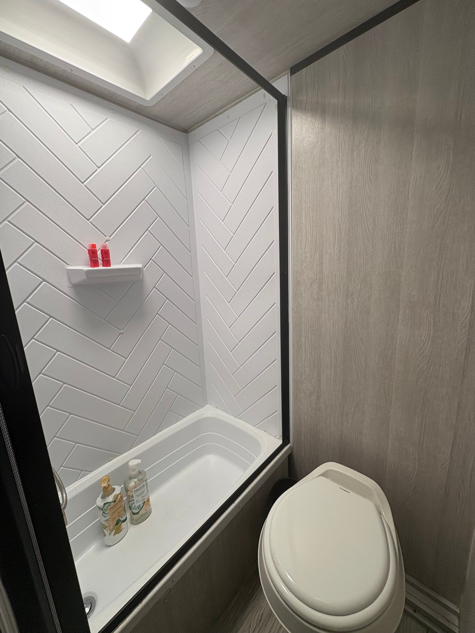 Clean RV rental bathroom with a white herringbone shower, bathtub, toilet, and wood-look walls. Features a skylight.