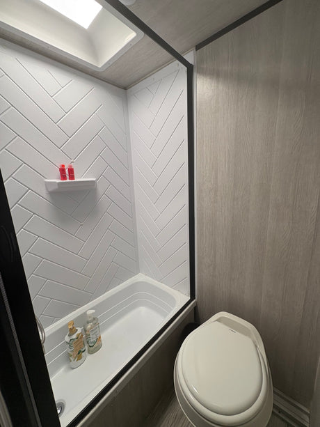 Clean RV rental bathroom with a white herringbone shower, bathtub, toilet, and wood-look walls. Features a skylight.