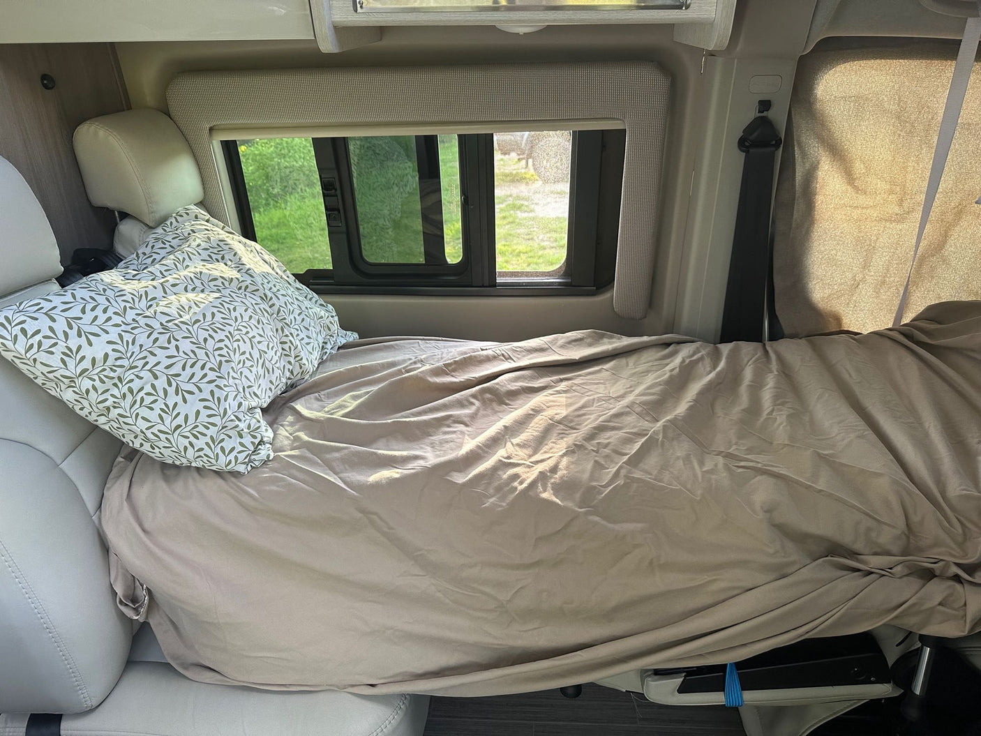 Cozy RV rental bed with a leafy pillow and beige blanket, offering a green view from the window.