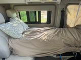 Cozy RV rental bed with a leafy pillow and beige blanket, offering a green view from the window.