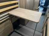 Inside an RV rental, a light-colored table extends into a compact living area with wooden bed slats and cabinets.