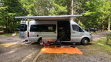A silver Mercedes-Benz Sprinter RV rental parked at a wooded campsite, featuring an extended awning, chairs, and mat.