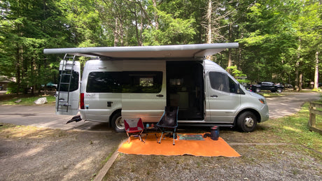 A silver Mercedes-Benz Sprinter RV rental parked at a wooded campsite, featuring an extended awning, chairs, and mat.