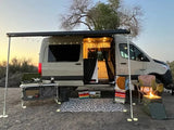 Cozy RV rental setup at sunset: camper van with awning, outdoor kitchen, chairs, & fire pit. Perfect van life camping!
