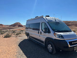 Silver rv rental camper van parked on a desert gravel road, with red rock hills under a bright blue sky.