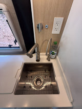 Modern RV rental kitchen: deep stainless steel sink with dual faucets, bright countertop, power outlet, & window view.
