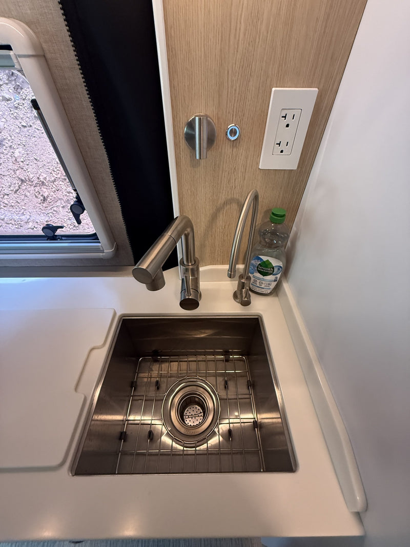 Modern RV rental kitchen: deep stainless steel sink with dual faucets, bright countertop, power outlet, & window view. — photo 16