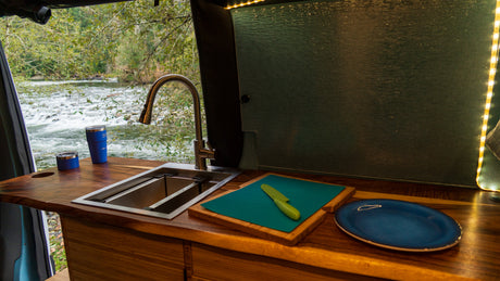 An RV rental kitchen setup with a sink, cutting board, and drinkware, offering picturesque views of a flowing river.